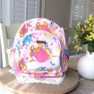 Handmade pink fairytale mini backpack with colorful storybook-inspired fabric displayed on a wooden table beside decorative flowers indoors.