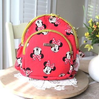 Handmade bright pink boutique mini backpack with playful character-inspired fabric displayed on a wooden table beside decorative flowers indoors.